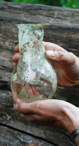 Überreste eines gläsernen Urinals, mit rundem Boden und gerade Hals. Gefäß ist an einie Stellen zerbrochen.
